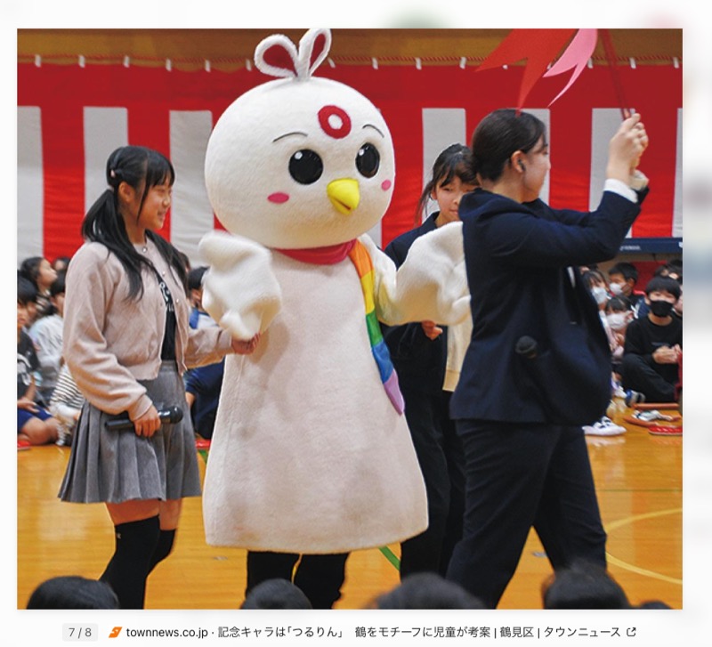 白い鳥の着ぐるみ。額に赤い輪、首に虹色のマフラー。体育館で児童に囲まれている